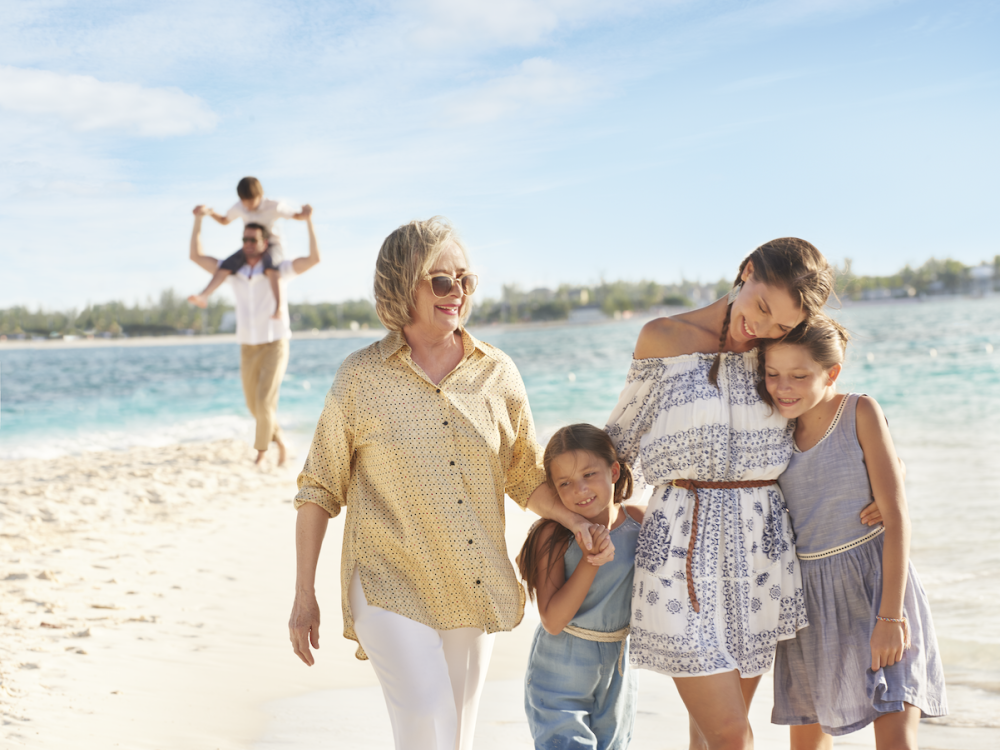 A grandma, mother, and two girls walk on a sandy beach with a dad and son in the background