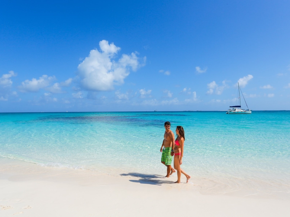 A couple walk hand in hand on the beach in Nassau Paradise Island and a boat is anchored in the distance. 