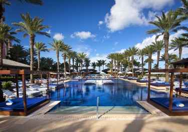 A couple relax on a large lounge chair by the pool on a sunny day.