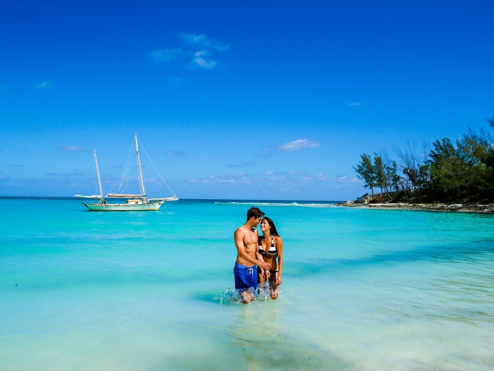 A couple in the turquoise waters of a private cove with a sailboat in the background.