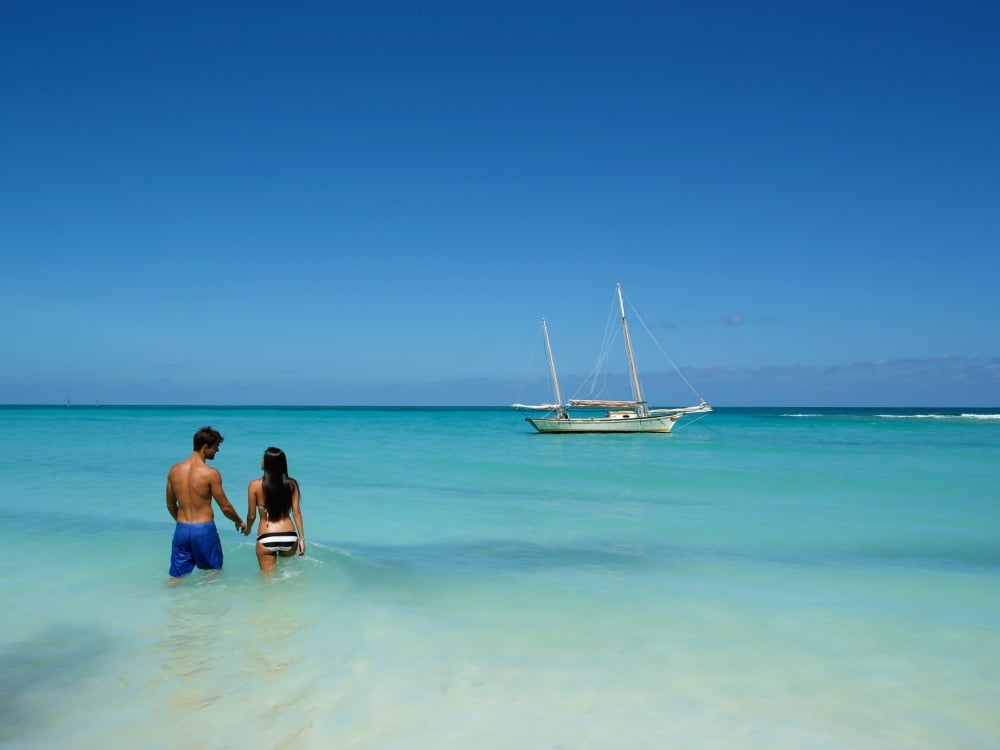 A couple holds hands while standing in the water looking at a sailboat in the distance.