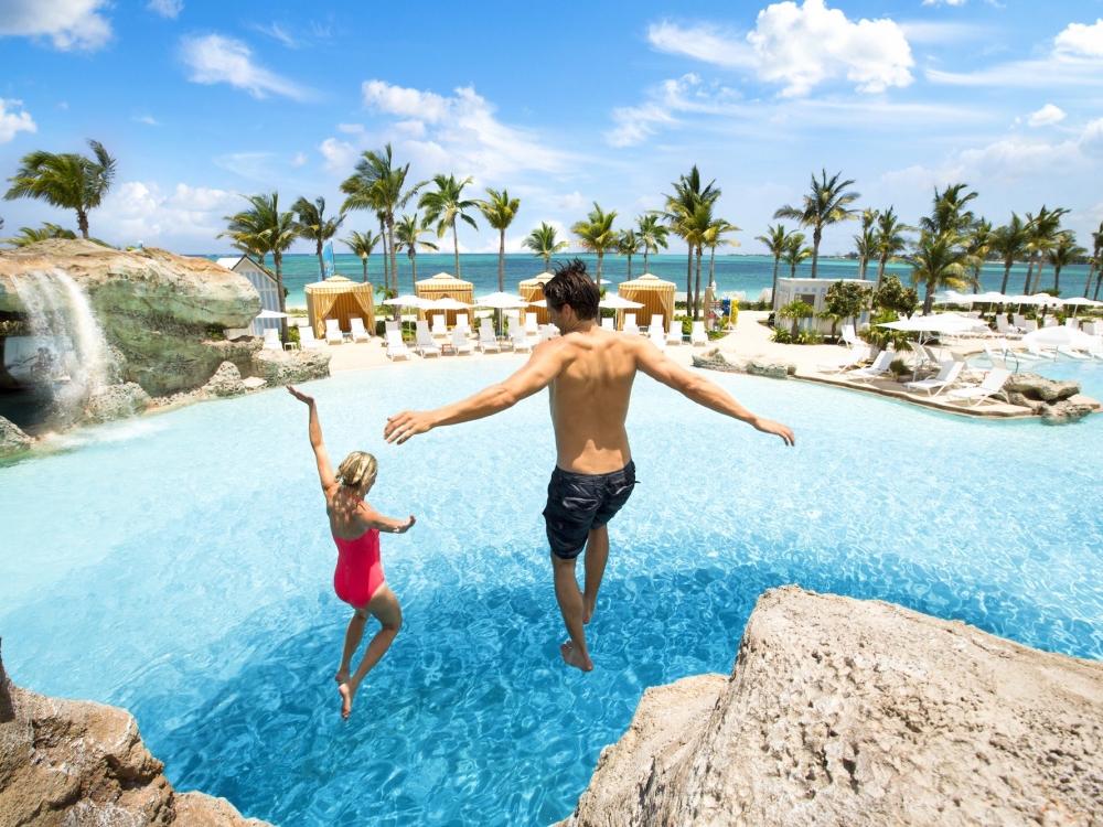 A young couple jumps off a cliff into a turquoise pool in The Bahamas. 
