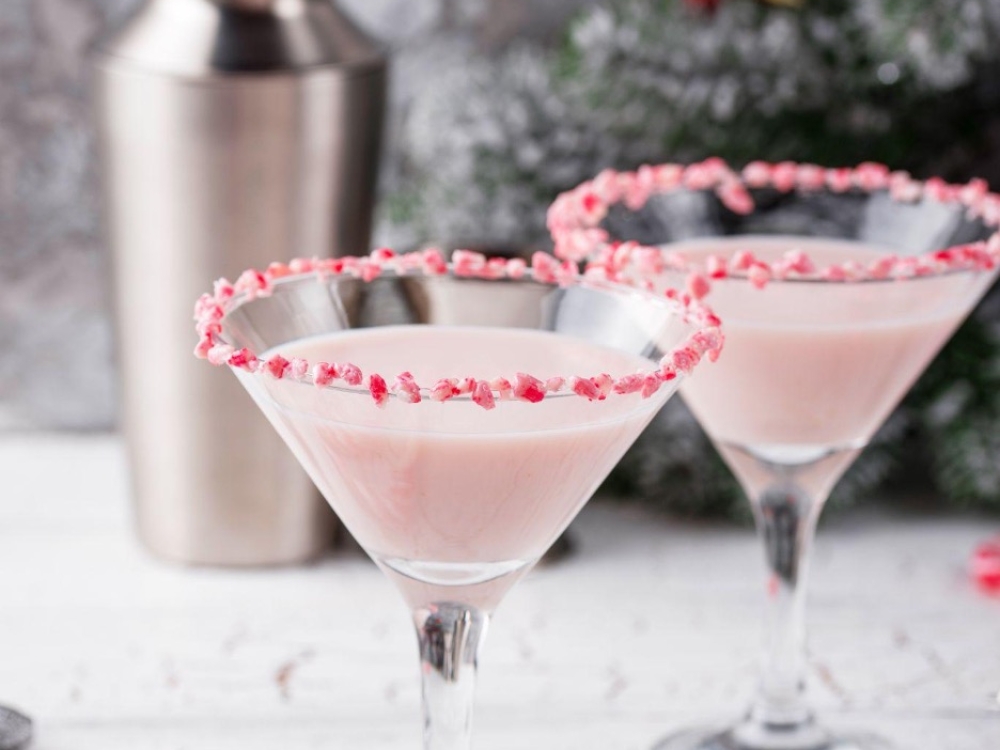 Two martini glasses with crushed candy cane on the rim and light pink liquid inside and a drink shaker in the background