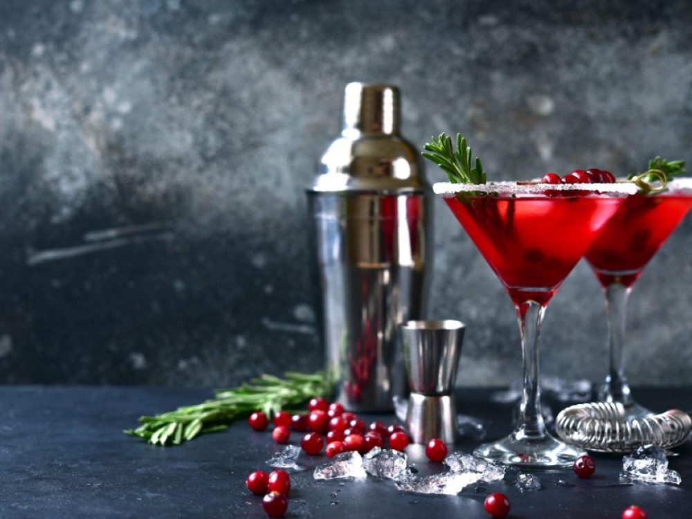 Two martini glasses filled with red liquid with cranberry garnish and a metal drink shaker and shot glass