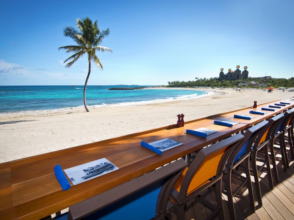 Wooden diner bar facing the white sand beach, turquoise waters, and palm tree of the beach at The Cove at Atlantis.