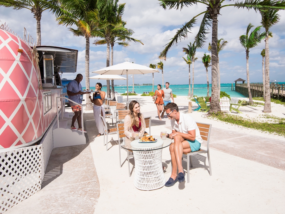 A server exits a pink beachside airstream to serve food to diners.