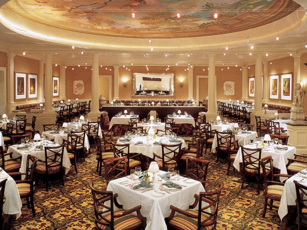Tables are set for dinner in a large dining area with white pillars and an intricately painted ceiling.