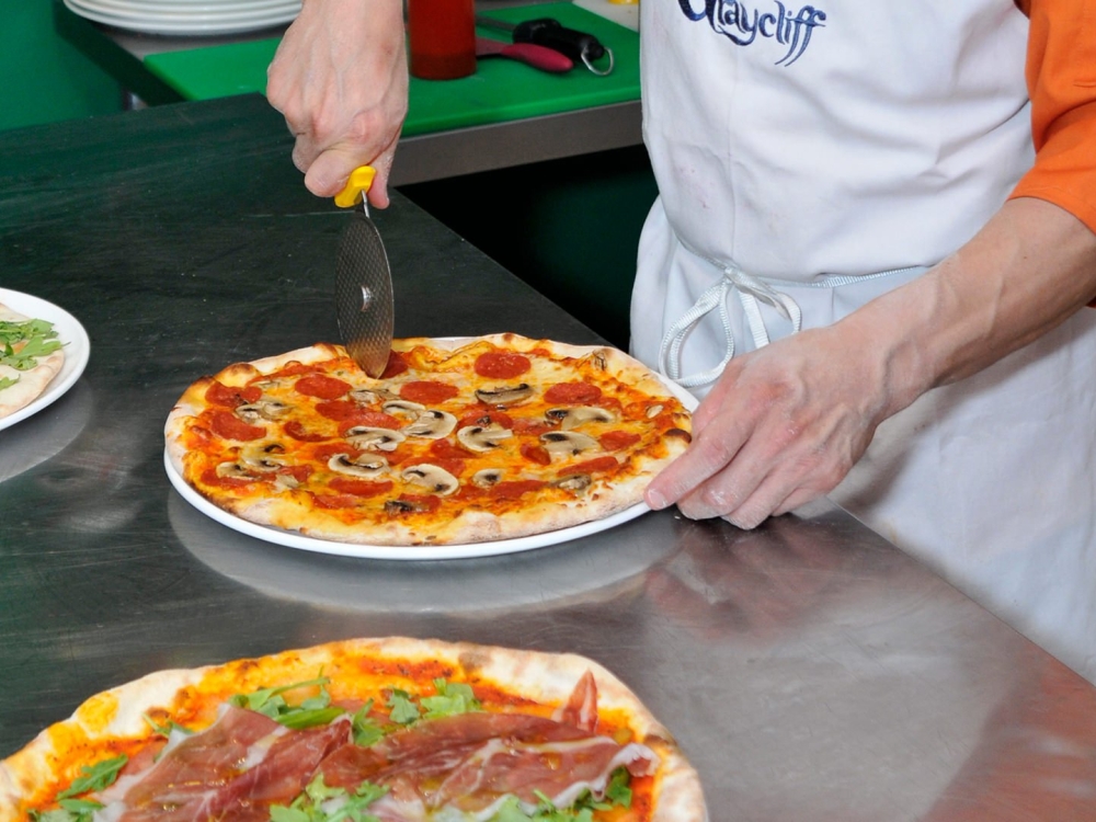 A man cutting a mushroom and pepperoni pizza with a pizza cutter over a stainless steel surface.