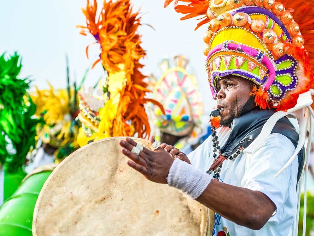 A man in a brightly colored headdress blowing a whistle and playing a drum at Carnival.