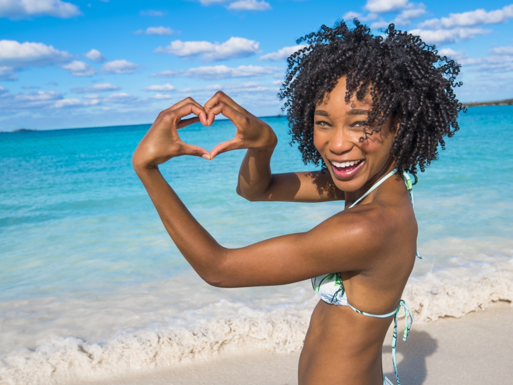 A woman smiles and makes a heart with her hands on the shores of Nassau Paradise Island. 
