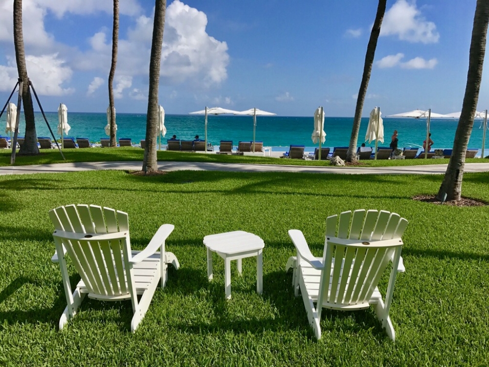 Two white chairs on a lawn overlooking an infinity pool and the ocean. 