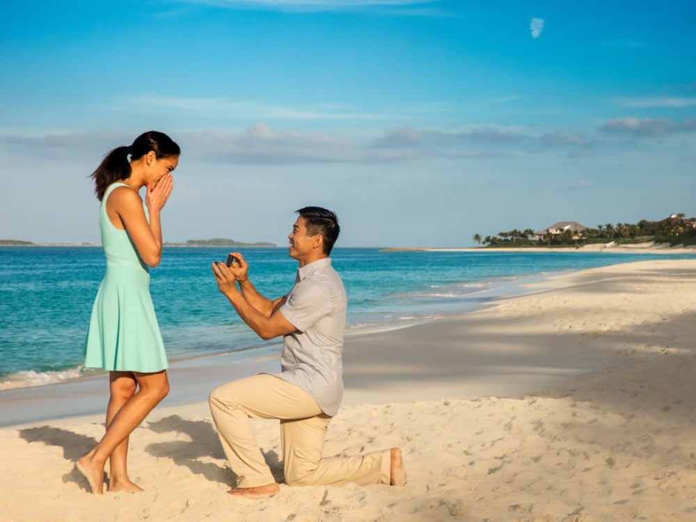 A man kneels on a Bahamas beach to propose to a woman.