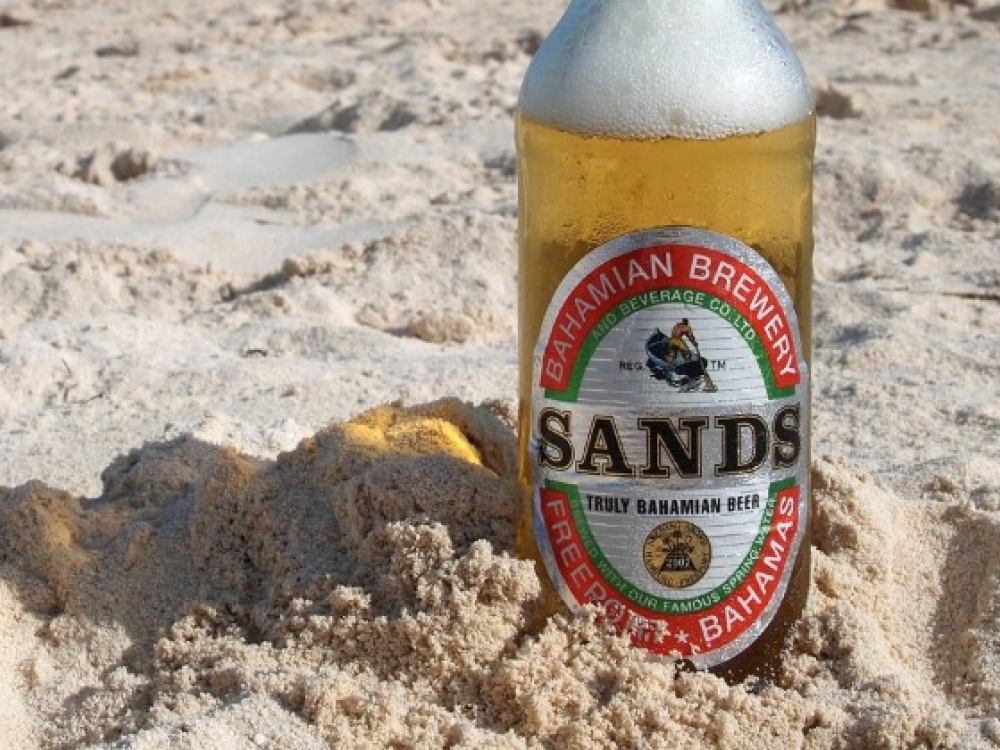 A bottle of Sands beer sticks out of the sand on a Bahamas beach. 
