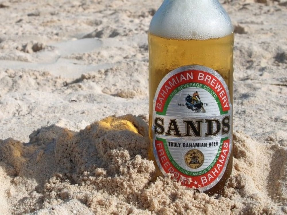A bottle of Sands beer sticks out of the sand on a Bahamas beach.