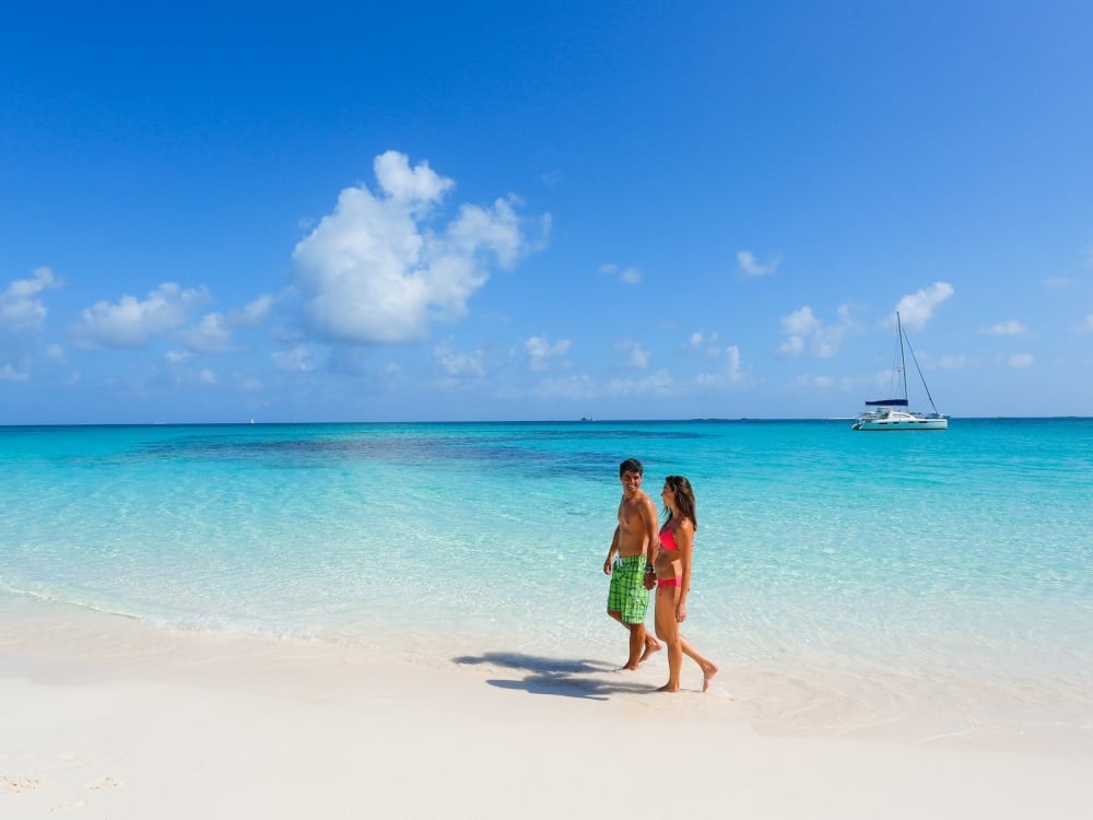 A couple walk hand in hand on the beach in Nassau Paradise Island and a boat is anchored in the distance.