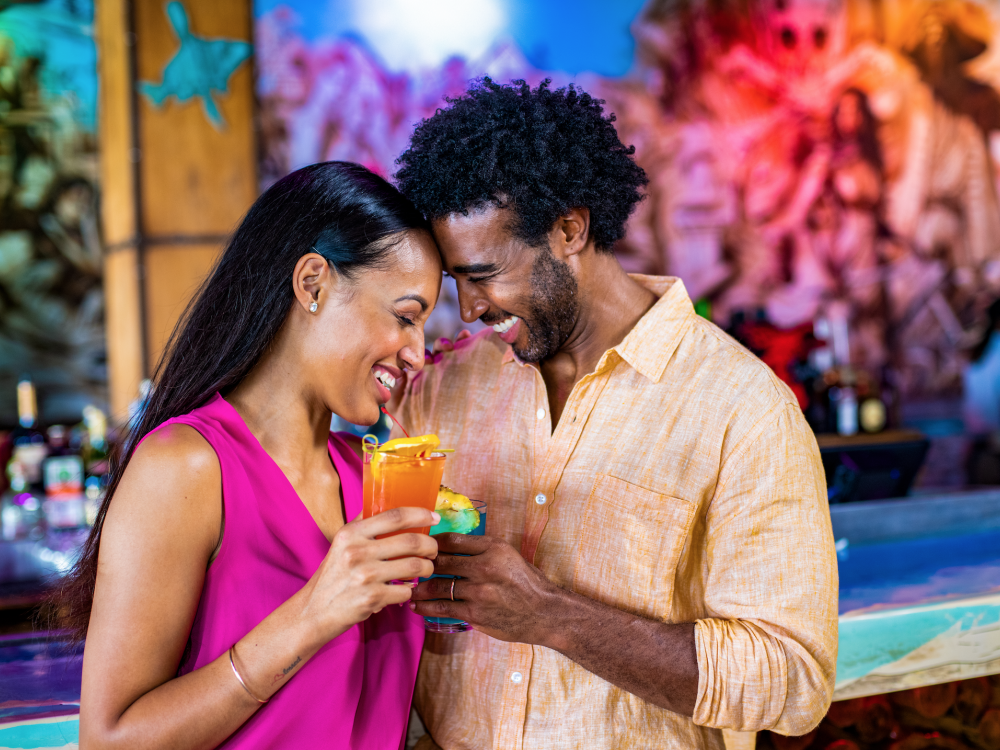 A couple toast with tropical cocktails against a colorful backdrop. 