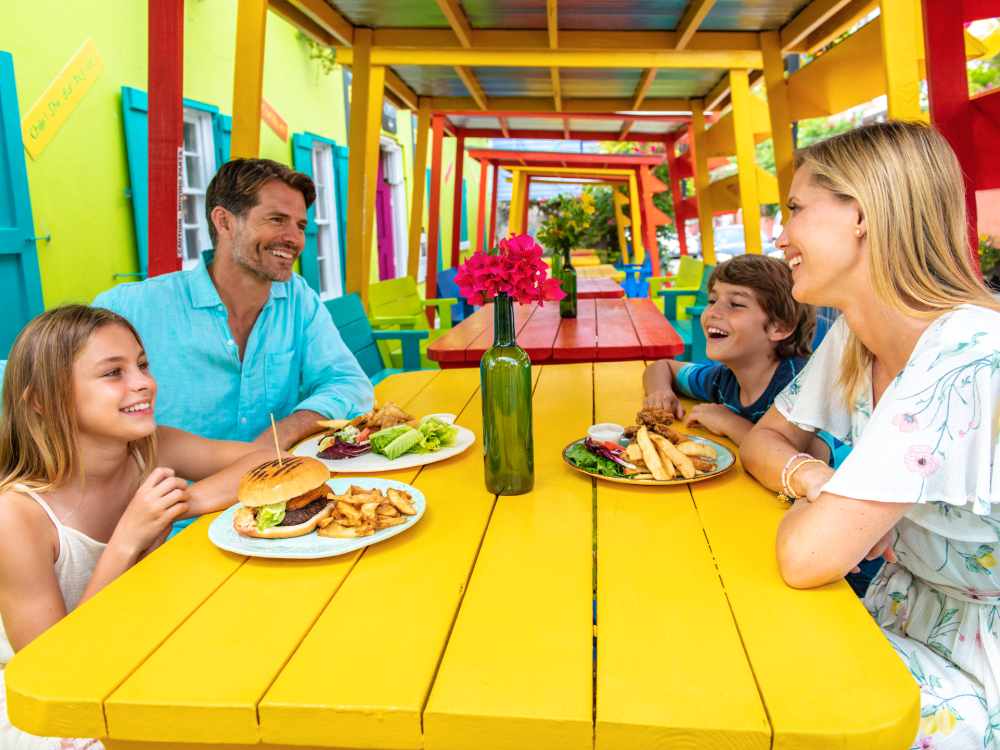 A family of four enjoys a casual lunch on a bright yellow picnic table. 