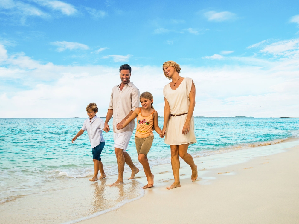 A family of four walking hand-in-hand down a tropical white sand beach.