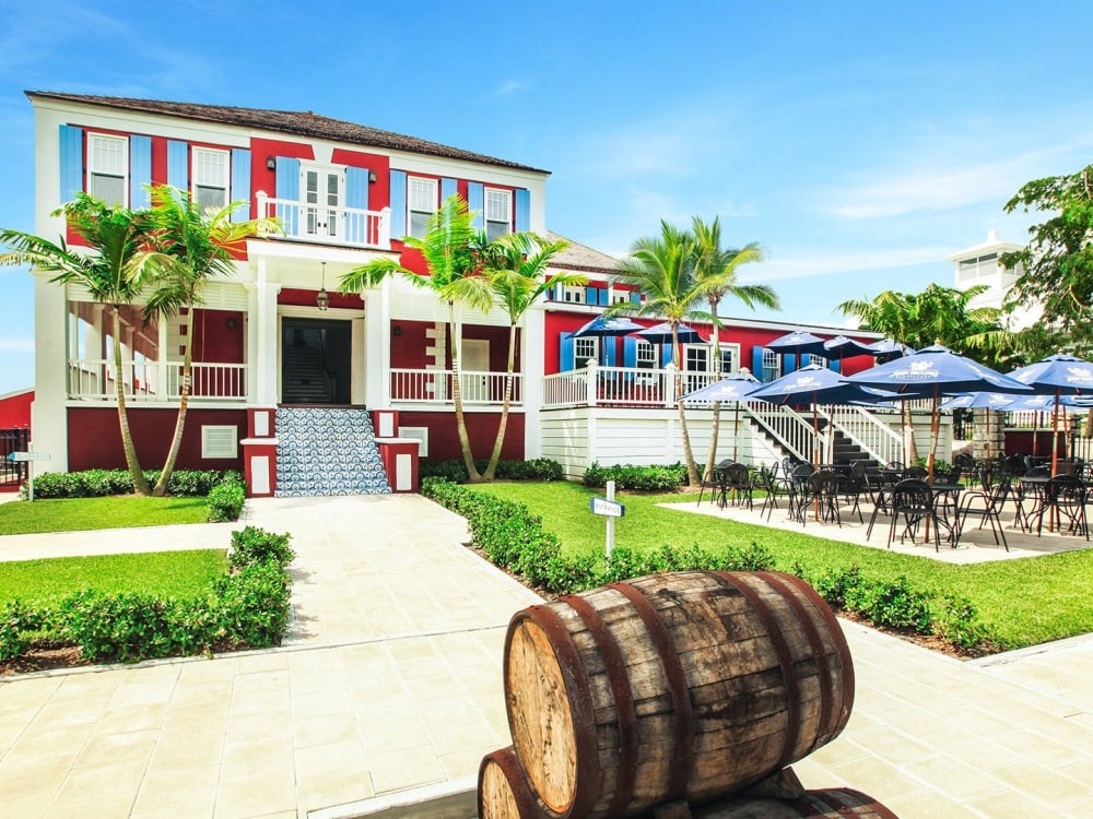 A wooden barrel sits outside of a large red house with patio tables and chairs outside.