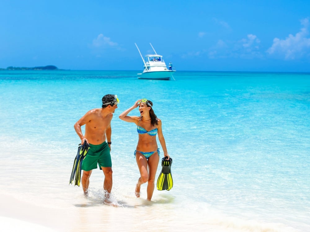 A man and woman walking out of the ocean together laughing and holding snorkelling gear.