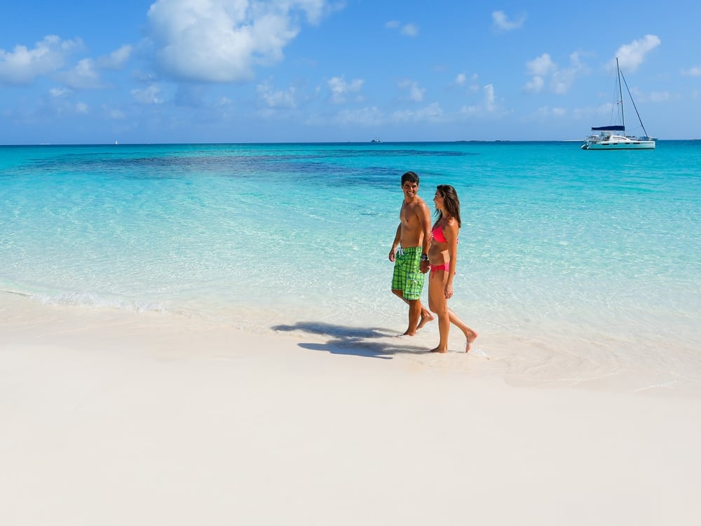 A man and a woman walk hand-in-hand down a white sand beach in Rose Island.