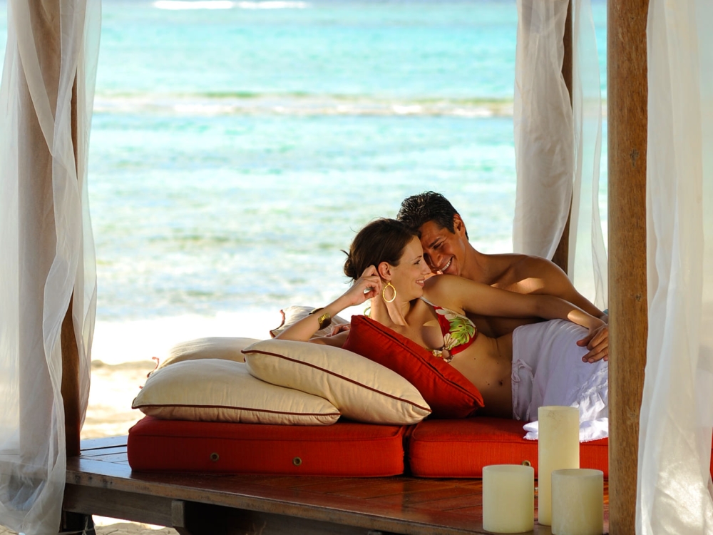 A man and woman lay oceanside together on a wooden pedestal filled with pillows and candles.
