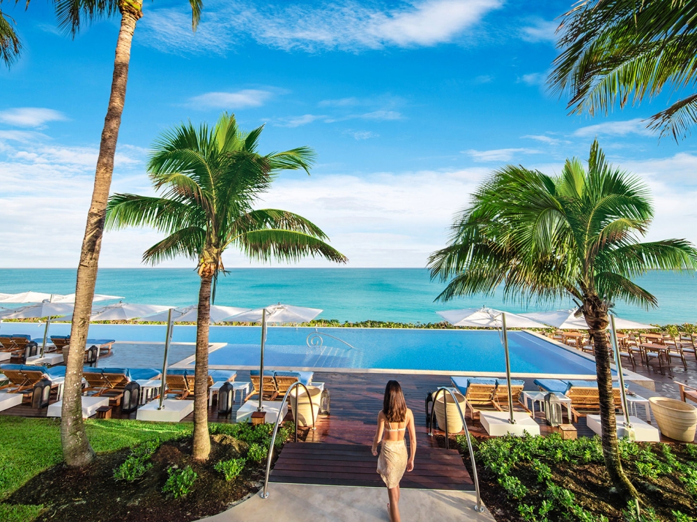 A woman walking towards a pool surrounded by palm trees and lounge chairs with a view of the ocean in the distance.