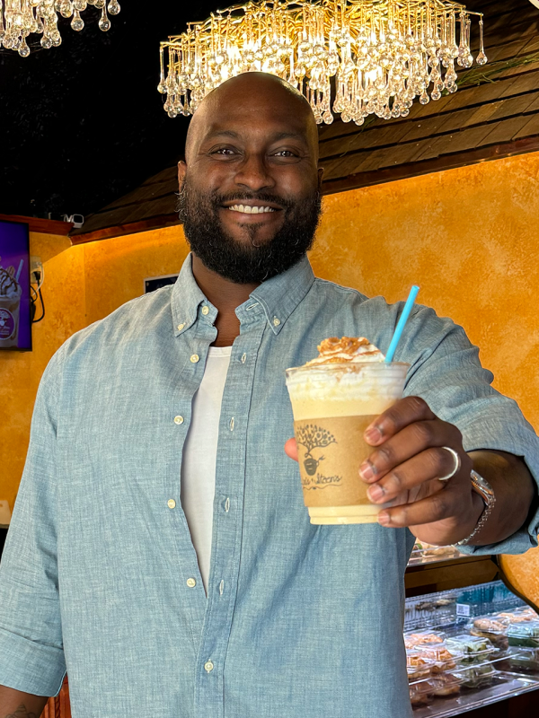 A man in a collared shirt holds a drink towards the camera smiling.