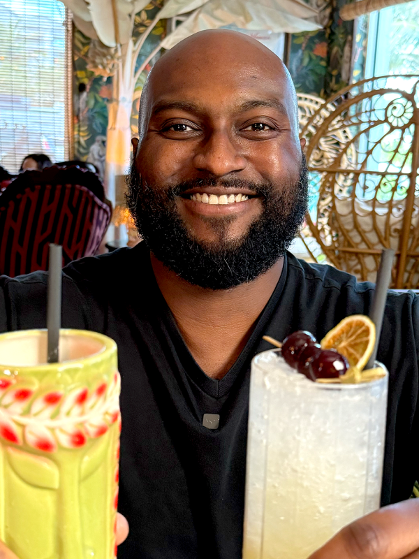 A man holds two cocktail glasses up to the camera and smiles.
