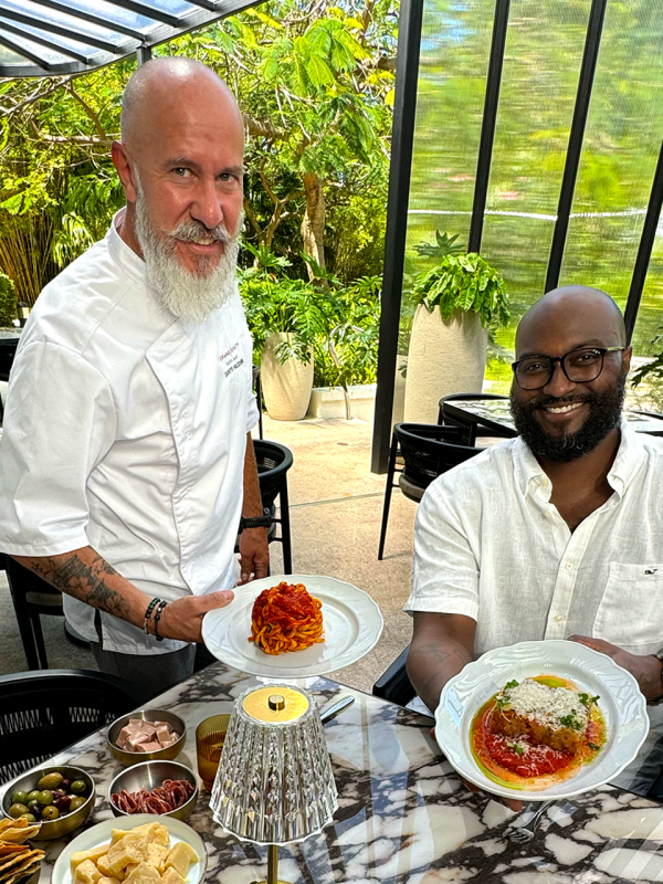 Two men, one sitting and one standing, hold up plates filled with food.