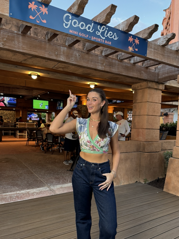 A woman in jeans and a crop top stands in front of a sign that says "Good Lies".