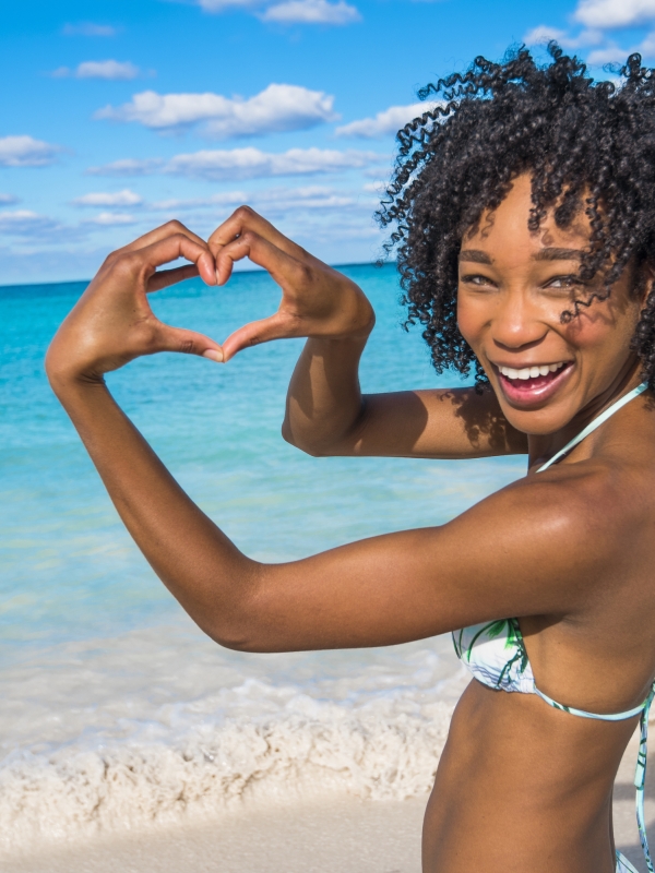 A woman smiles and makes a heart with her hands on the shores of Nassau Paradise Island. 
