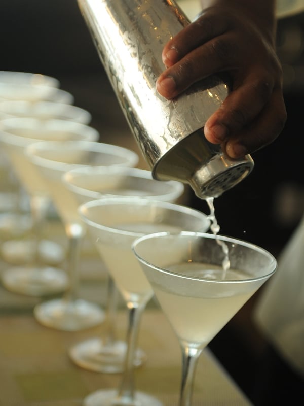 A freshly made cocktail is poured from a cocktail shaker into a row of martini glasses. 