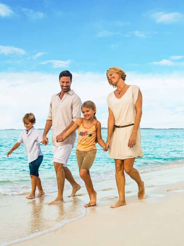 A family of four walking hand-in-hand down a tropical white sand beach.