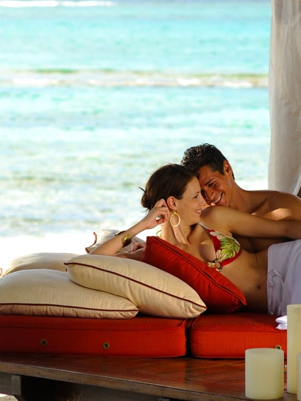 A man and woman lay oceanside together on a wooden pedestal filled with pillows and candles.