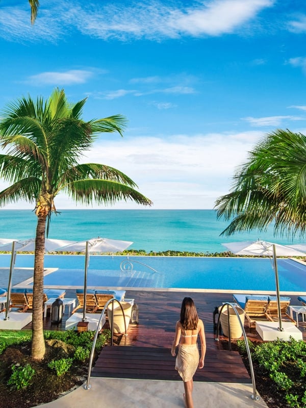 A woman walking towards a pool surrounded by palm trees and lounge chairs with a view of the ocean in the distance.